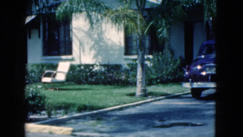1975 Florida residential exterior with palm trees, lawn chair, shrubs, curb, and a dark parked car partly visible at the right edge.