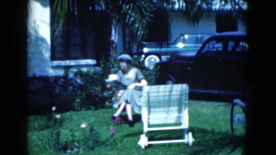 1975 Florida home-movie frame of a woman seated outdoors reading a newspaper on a front lawn, with parked cars, palms, and a house behind her.