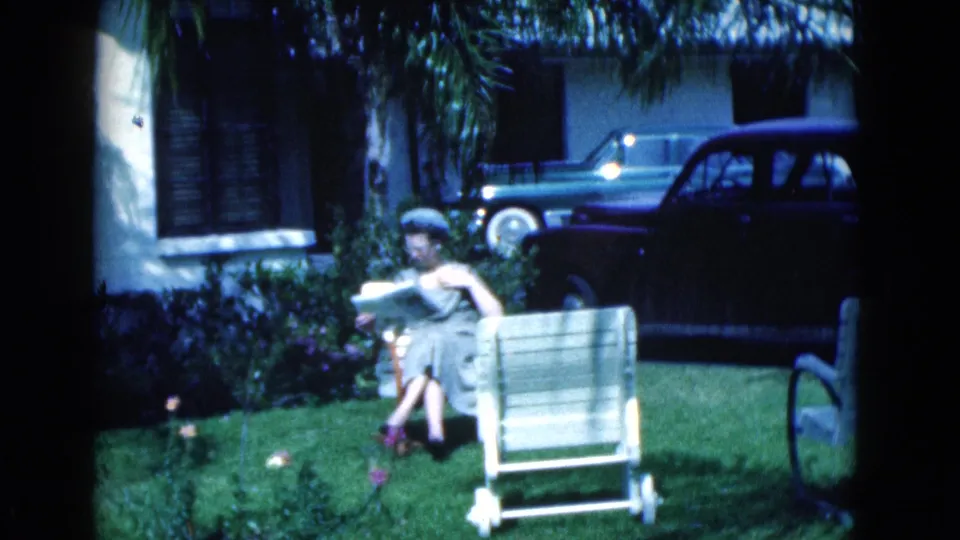 1975 Florida 8mm frame showing a woman reading on a front lawn near an empty chair, with residential facade, palms, and parked cars behind.