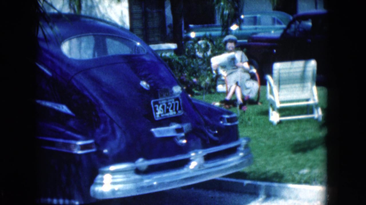 Woman seated on a lawn chair reading a newspaper beside an empty chair and parked cars.
