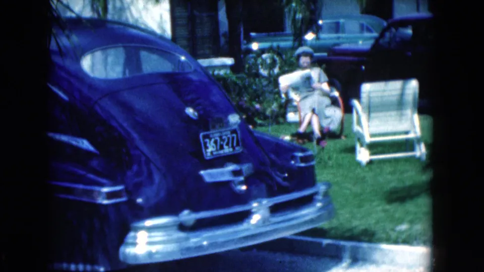 1975 Florida home-movie frame showing a woman reading a newspaper on a lawn near an empty chair and vintage parked cars.