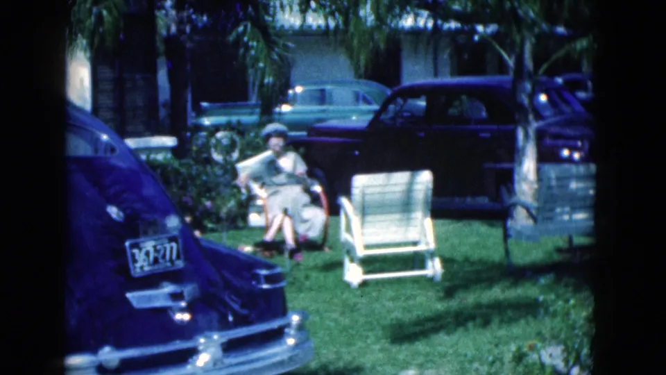 Home-movie frame from 1975 Florida showing a woman reading in a suburban lawn setting with classic cars nearby.