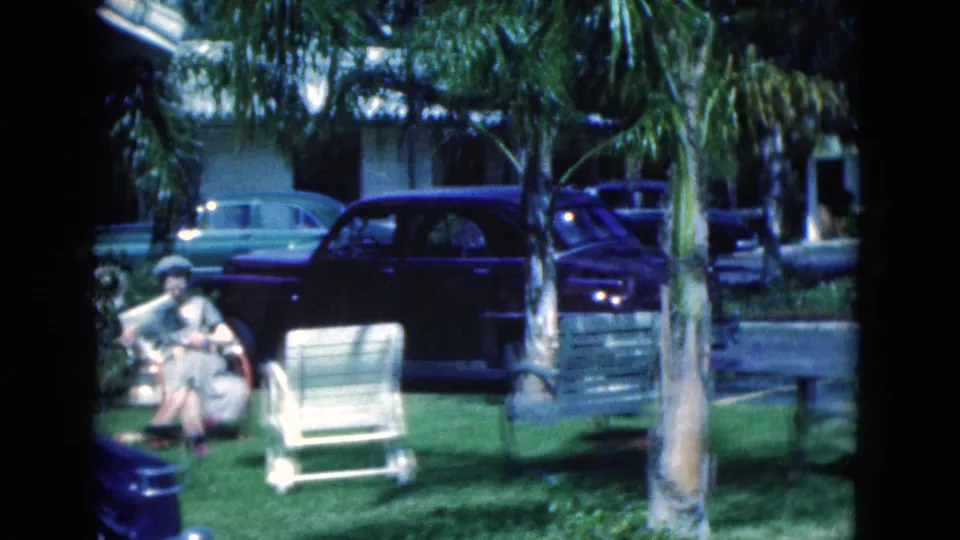 Wider 1975 Florida yard view with a woman reading on the grass, an empty lawn chair, palm tree, and parked automobiles.