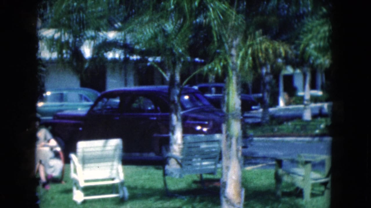 1975 Florida 8mm frame showing a seated woman on a lawn, palm trees, benches, and parked cars outside low cabin-like buildings.