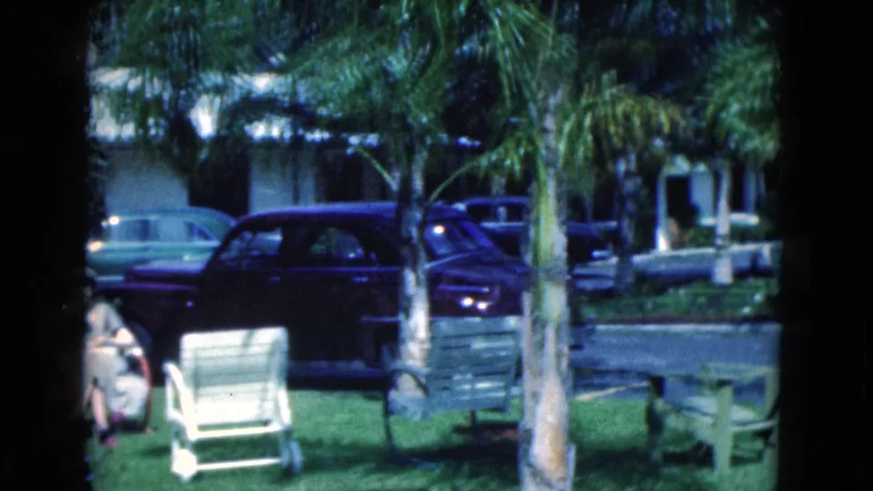 1975 Florida 8mm frame showing a seated woman on a lawn, palm trees, benches, and parked cars outside low cabin-like buildings.