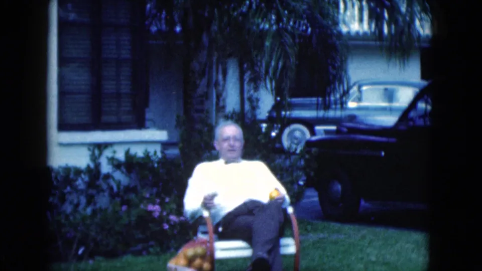 Older man sits on a lawn chair in a Florida yard, with house windows, shrubs, grass, and parked vintage cars behind him.