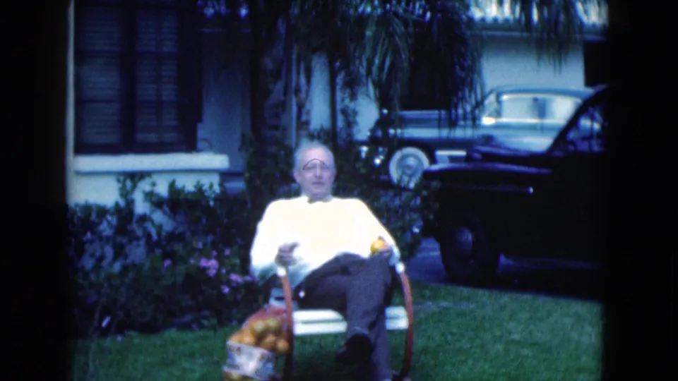 1975 Florida home-movie frame of an older man seated outdoors on a lawn chair, holding a bag of oranges near parked cars and a house.