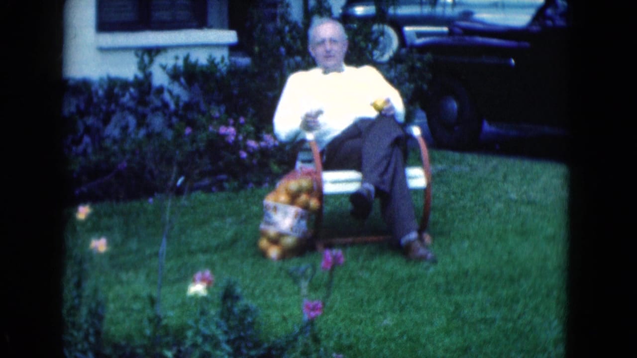 1975 Florida frame showing the seated man holding fruit while a bag of oranges rests next to the chair on the grass.
