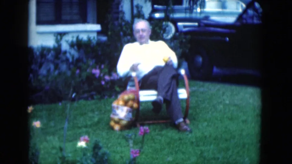 Soft 8mm frame of an elderly man seated in a Florida yard, with flowers in the foreground and a bag of oranges by his chair.