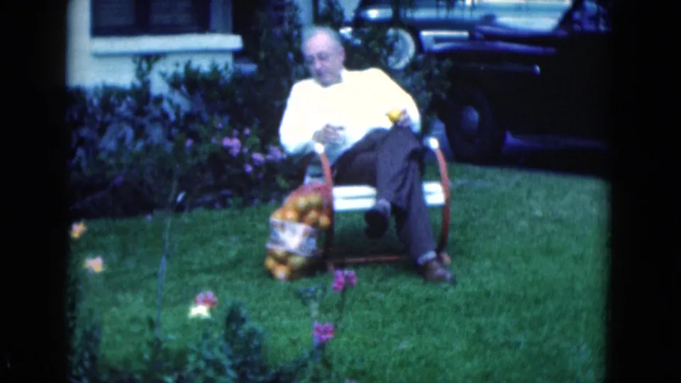 1975 Florida home-movie frame with the seated man looking down near a bag of oranges, lawn flowers, house facade, and cars behind him.
