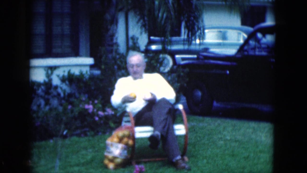 Older man holds an orange while seated in a chair on a lawn beside a full bag of oranges.