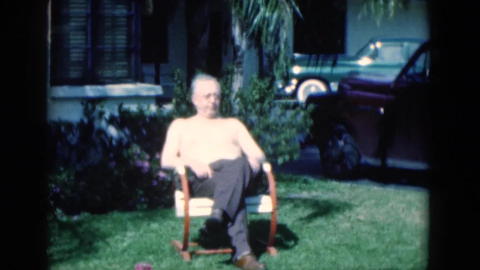 1975 Florida 8mm frame of an elderly man looking down from a lawn chair, with a suburban yard and cars behind him.