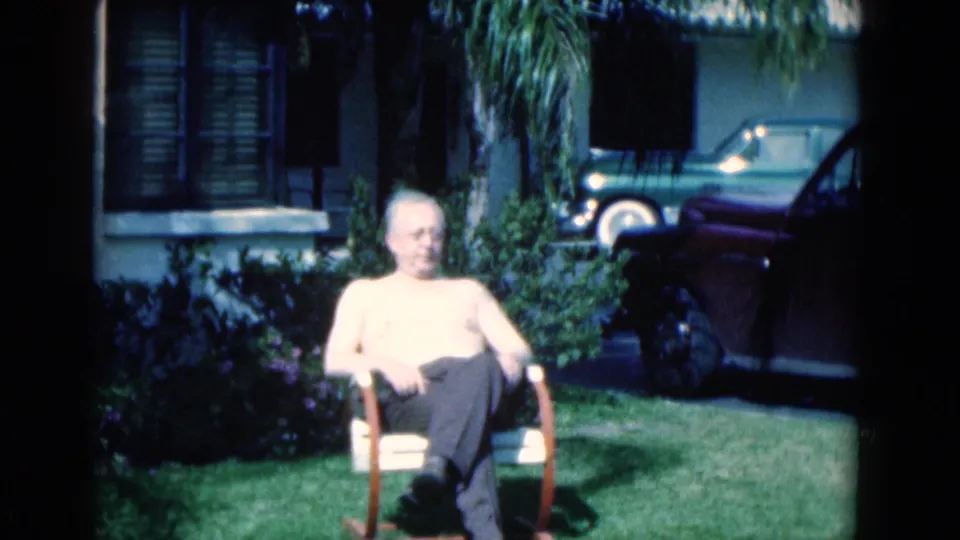 1975 Florida home-movie frame of an elderly man seated in a sunny suburban yard with palms, shrubs, and parked cars nearby.