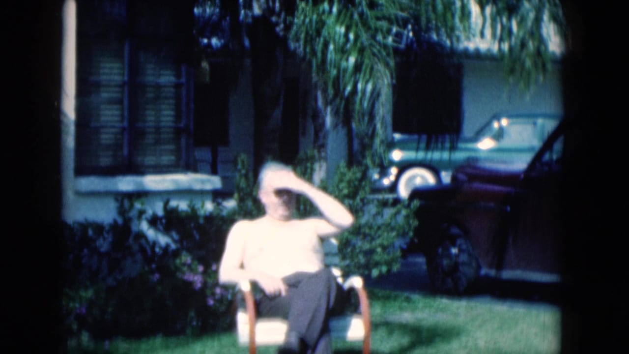 Man seated on a lawn chair in bright sun near a house and parked car.