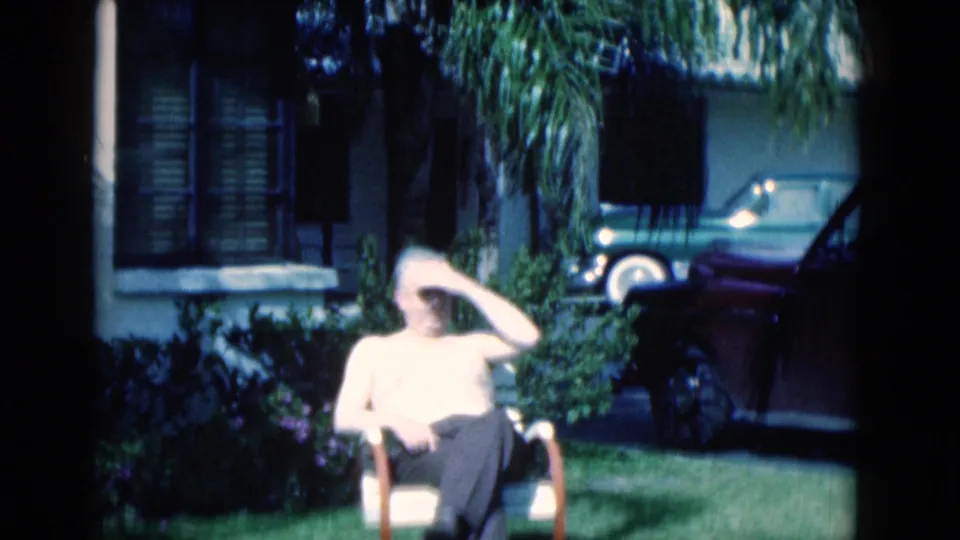 1975 Florida 8mm frame of a man seated on grass, shading his eyes, with a house, plantings, and parked car behind him.