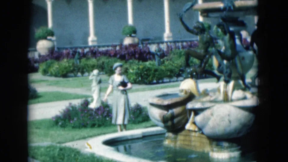 1975 Florida 8mm frame of a woman in a dress and hat beside a formal garden fountain with statues, hedges, and flower beds.