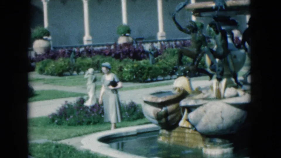 1975 Florida home-movie frame showing a woman by a sculptural garden fountain, with clipped hedges, flowers, and a building backdrop.