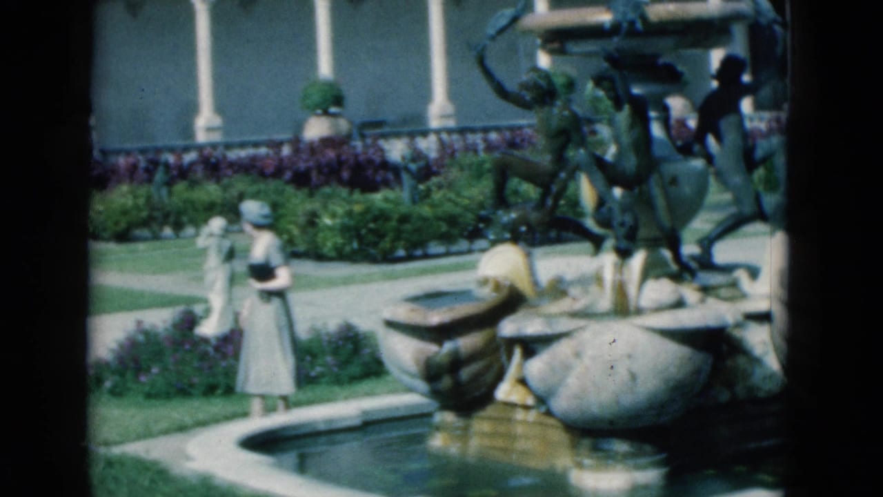 Woman in a light dress and hat walks near an ornate fountain in a landscaped courtyard.