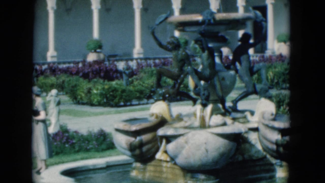 A woman stands near the edge of a courtyard fountain with arches and planting beds behind.