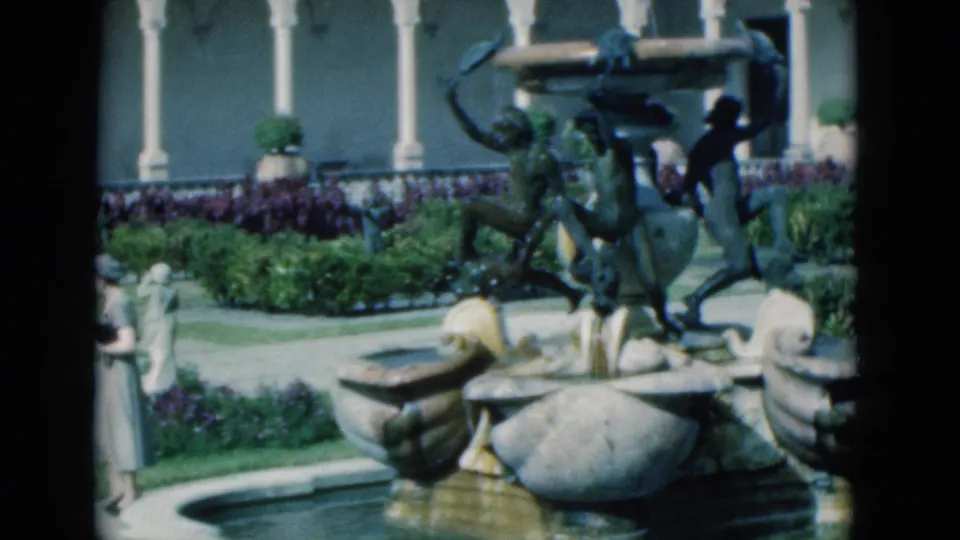 The woman appears near the fountain edge as bronze figures, stone basins, garden beds, and white colonnade arches fill the courtyard view.