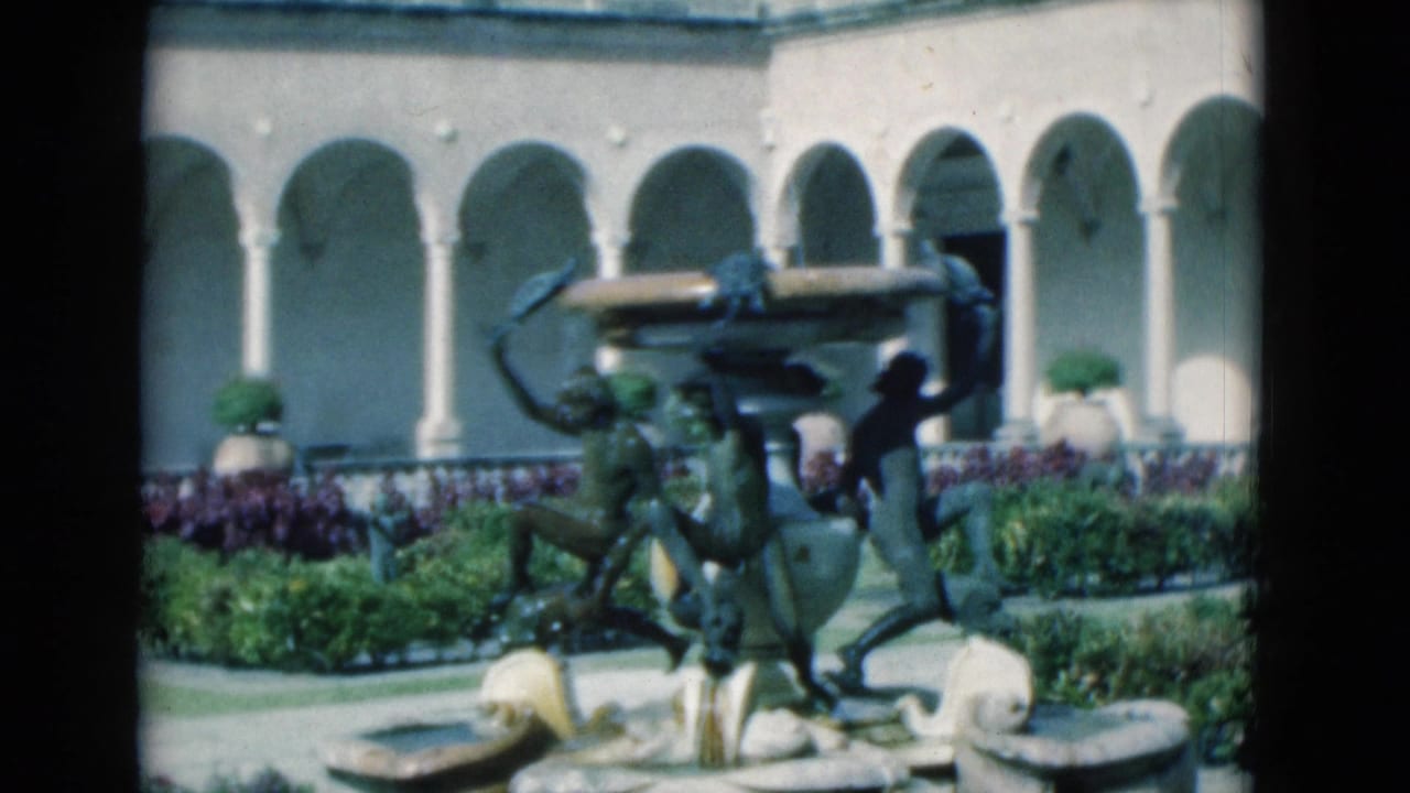 Ornate courtyard fountain with bronze figures in front of arched colonnades and flower beds.