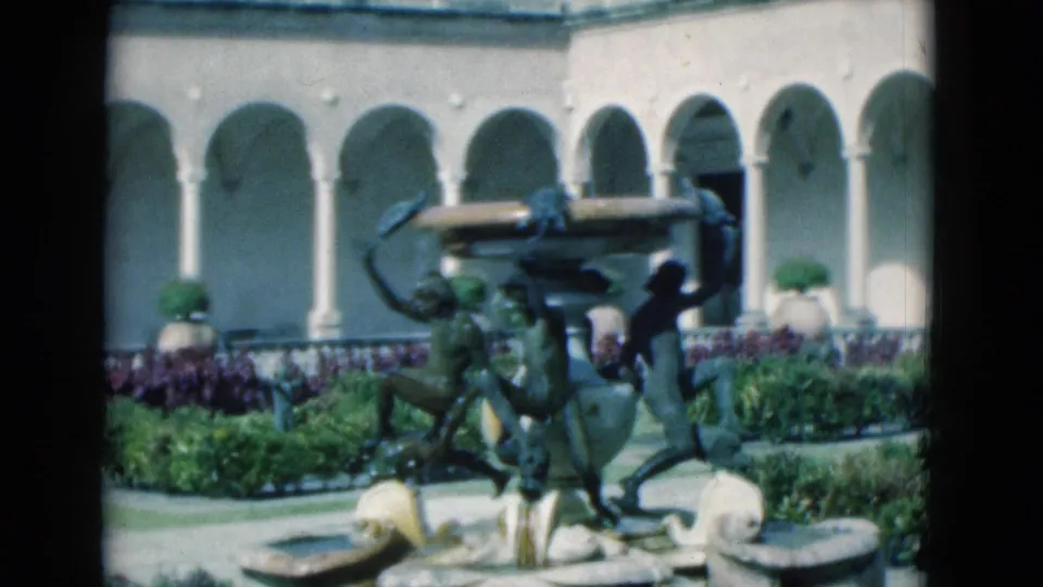 1975 Florida 8mm frame showing an ornate fountain with bronze figures, shell basins, trimmed greenery, flowers, and arched colonnades.