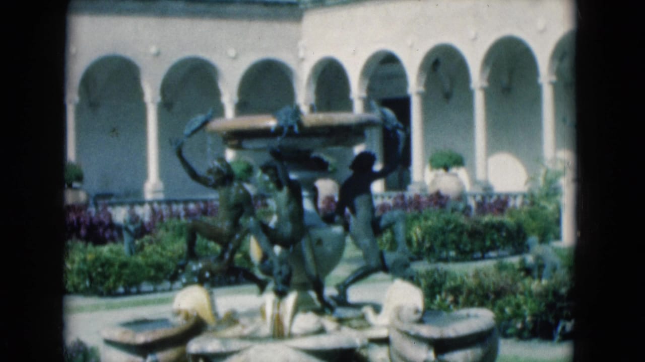 1975 Florida home-movie frame of the courtyard fountain, with bronze figures, water basins, flower beds, and white arches in the background.