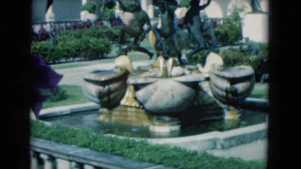 1975 Florida 8mm frame showing the fountain's lower basins, central sculpture, water, greenery, and a partial visitor at the left edge.