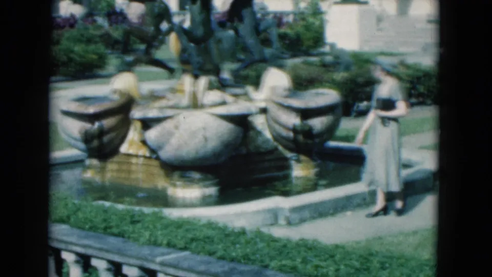 1975 Florida 8mm frame of a woman in a light dress and hat standing near an ornate fountain with greenery and arched architecture behind her.