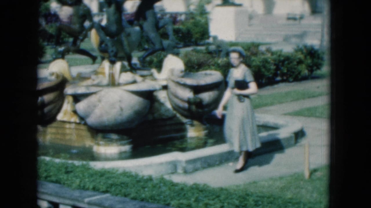 Woman in a light dress and hat standing beside an ornate garden fountain.
