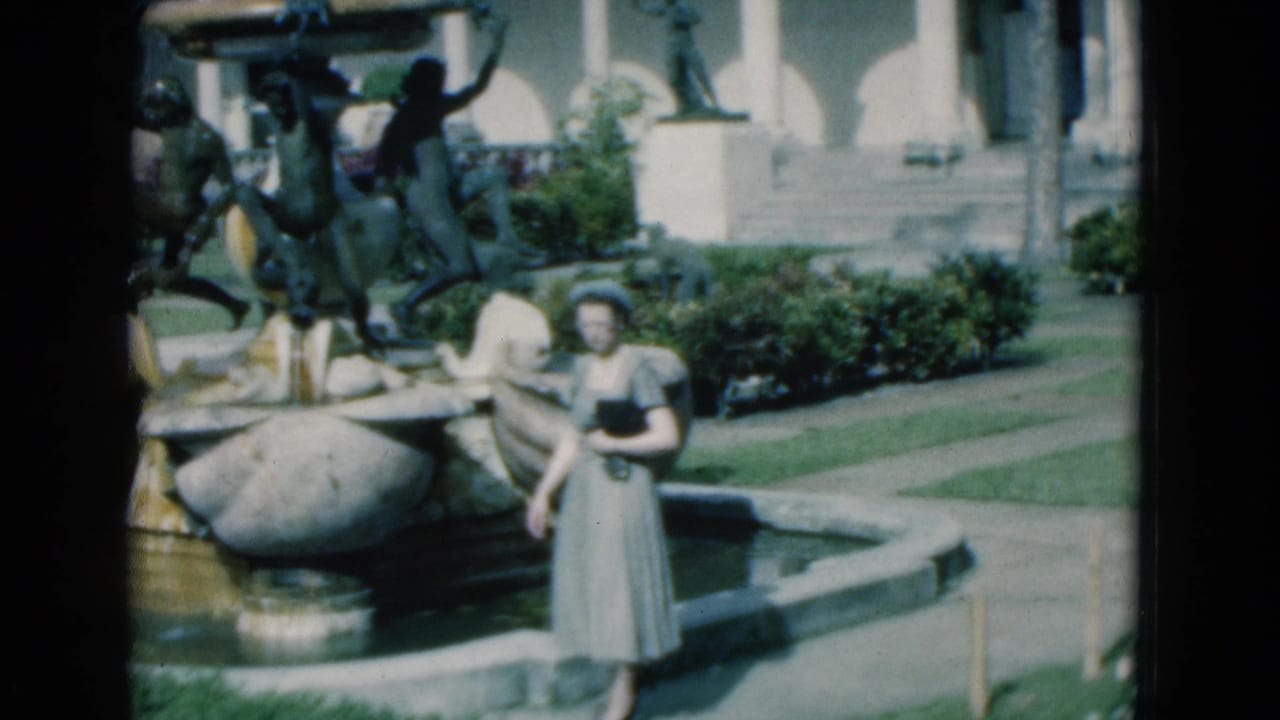 Woman walking near a large fountain with sculptures and garden paths.