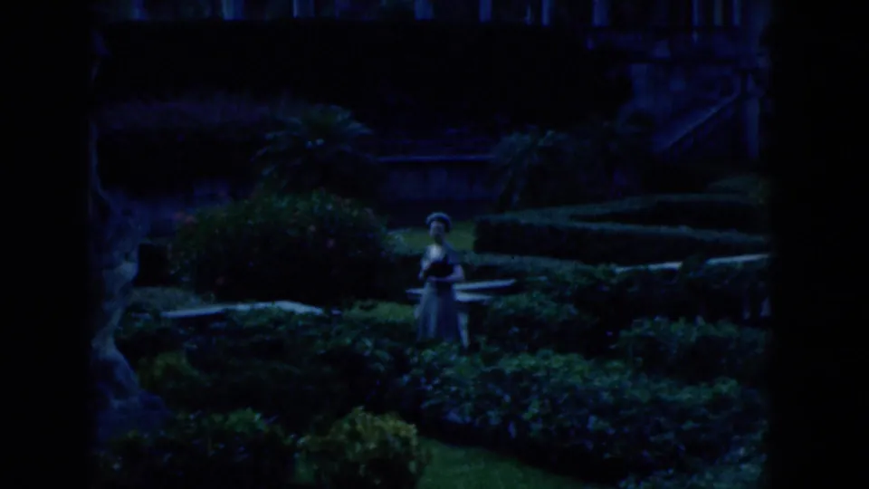 1975 Florida 8mm frame showing the woman farther back in shaded ornamental gardens with hedges, planting beds, and stonework nearby.