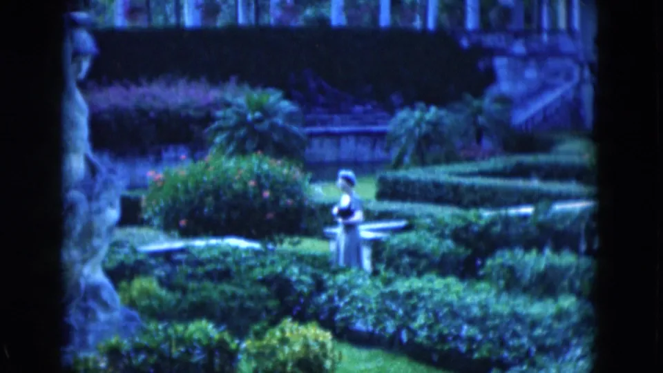 1975 Florida home-movie frame with the woman in profile among trimmed hedges, bright greenery, palms, and garden paths.
