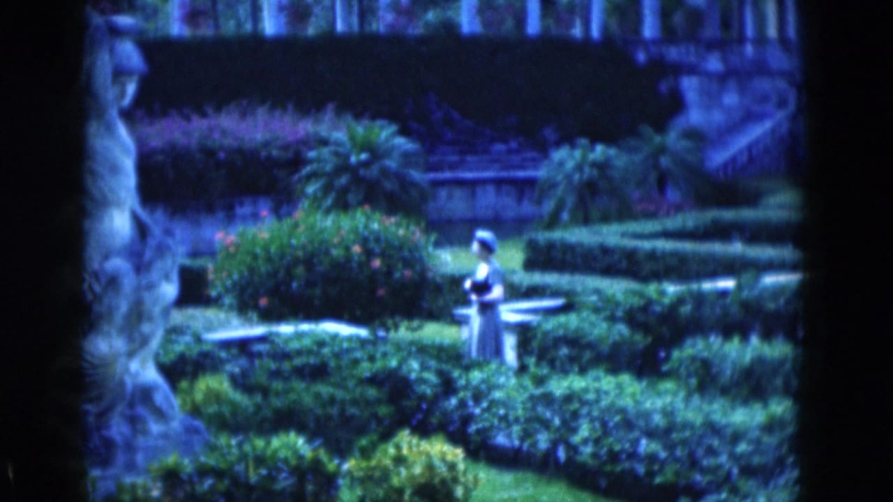 Woman in a light dress and hat stands among hedges near a stone statue and garden bench.
