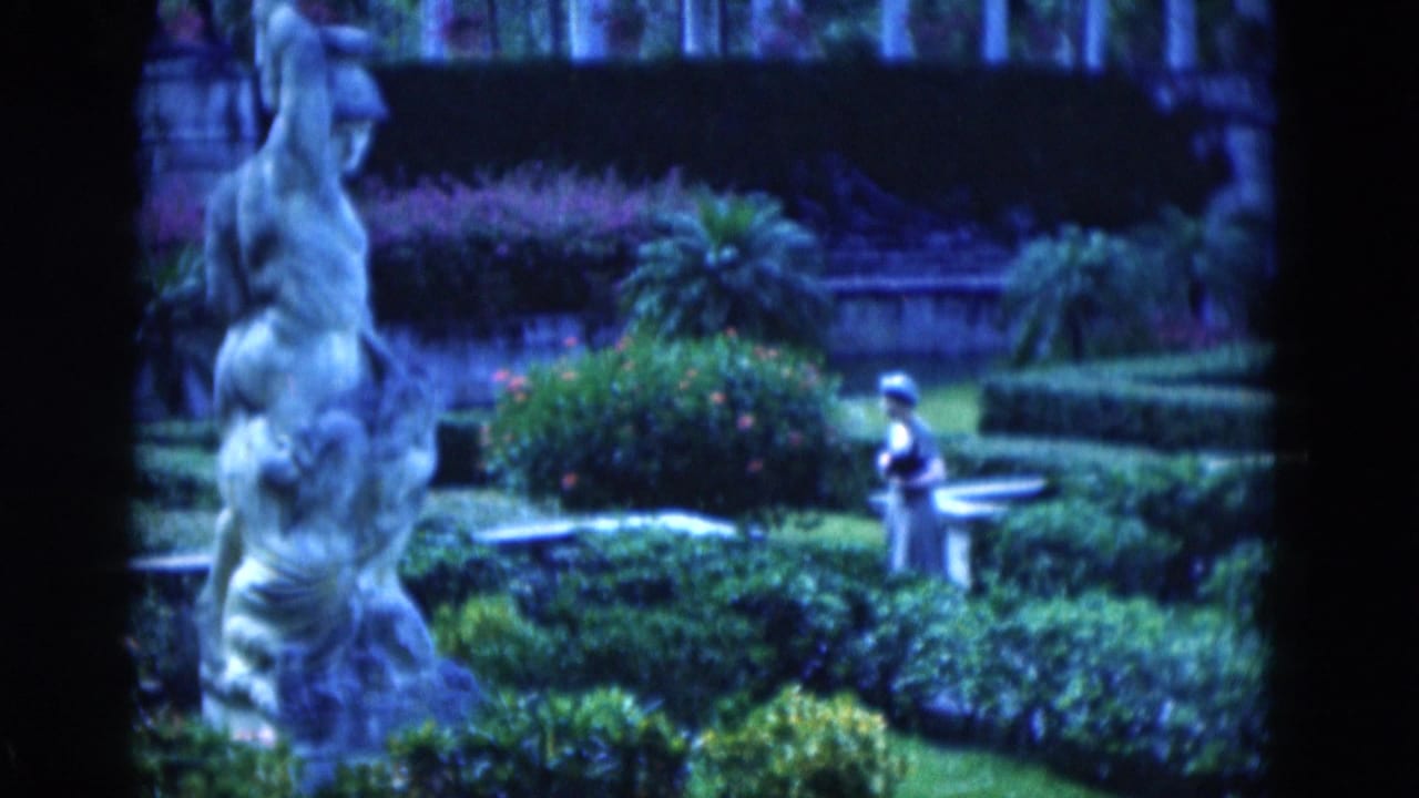 Woman stands near a stone bench in a formal garden with hedges and a tall statue in the foreground.