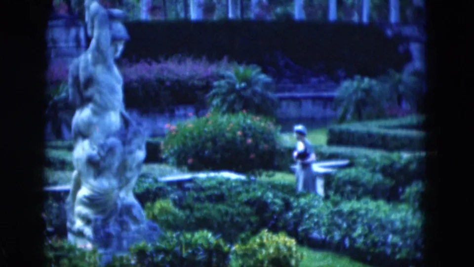 A 1975 Florida garden scene shows a woman near a bench, framed by clipped hedges, flowering shrubs, and a large classical statue in front.
