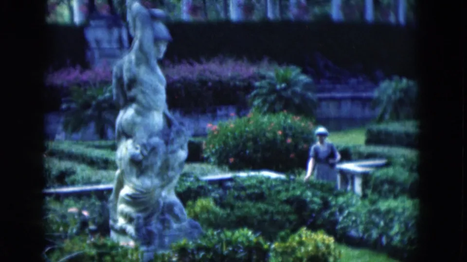 The 8mm frame places a classical stone statue in the foreground while a woman moves behind hedges near a bench in a Florida garden.