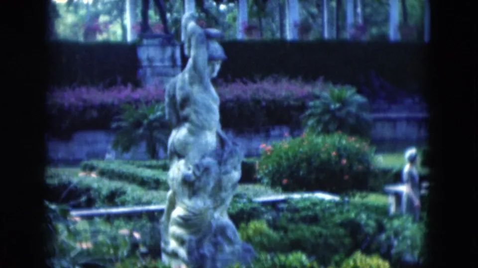 A 1975 Florida garden view centers a tall classical statue with clipped hedges and flowering shrubs, with the woman partly visible near the right edge.
