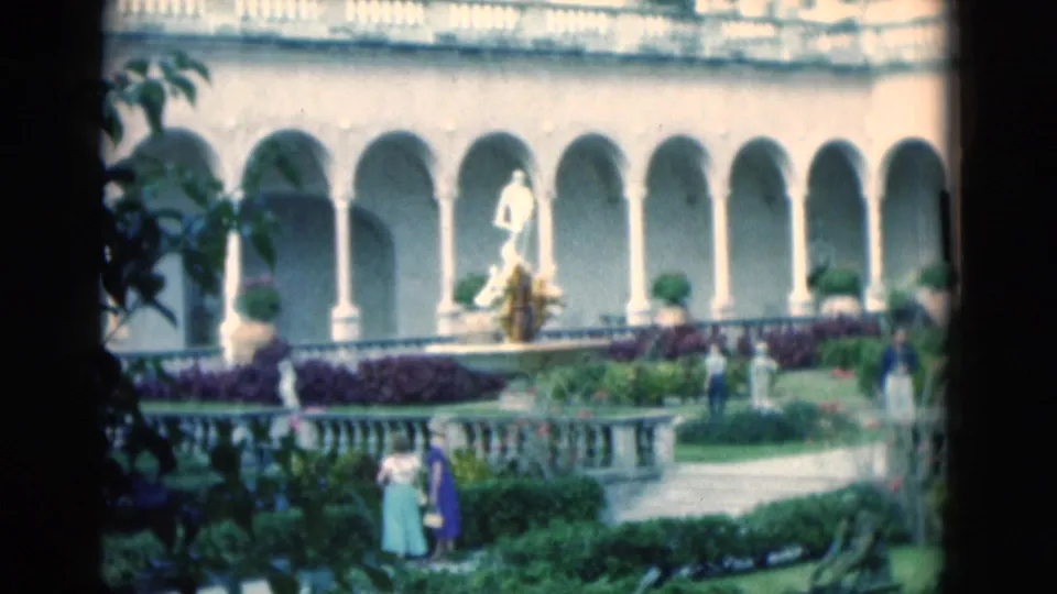 1975 Florida 8mm view of a grand arched colonnade, formal gardens, a fountain statue, and visitors walking below.