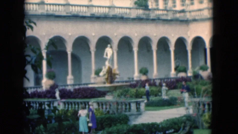 1975 Florida home-movie frame showing a formal courtyard with arched colonnade, statue, paths, hedges, and distant visitors.