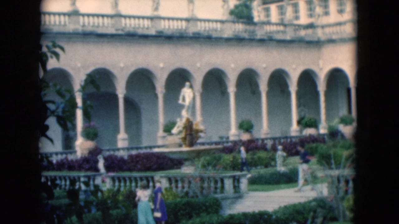 1975 Florida 8mm frame of a columned courtyard garden with statues, balustrades, shrubs, and distant visitors.