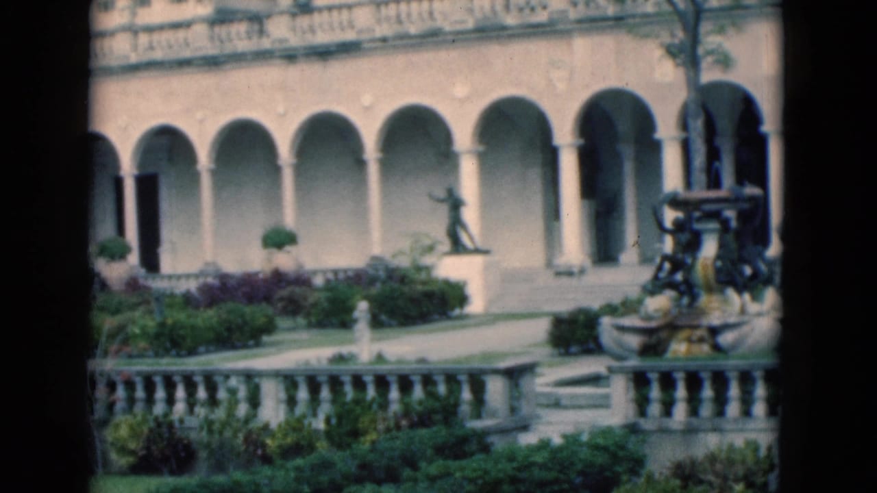 Arched building facade behind a garden with statues, shrubs, and a fountain at right.