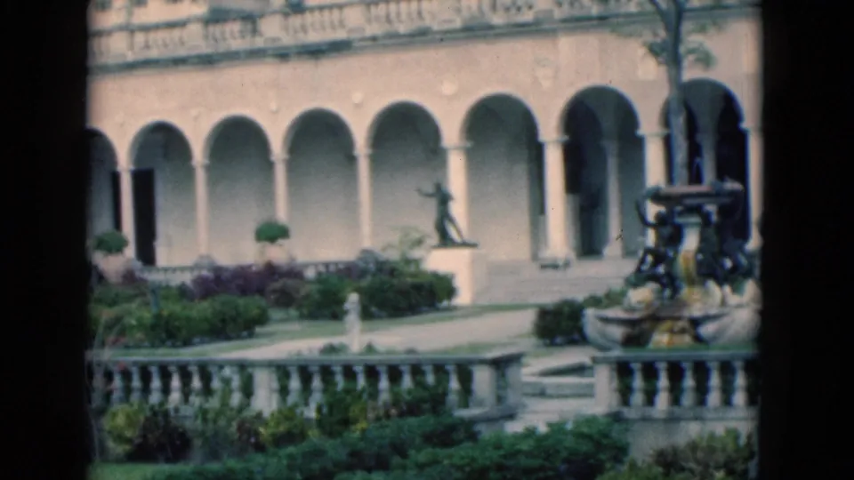 1975 Florida view across a landscaped courtyard toward arches, a pedestal statue, trimmed beds, and an ornate fountain.