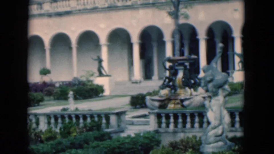1975 Florida 8mm frame showing a formal garden courtyard with colonnades, fountain sculpture, balustrade, and dense greenery.