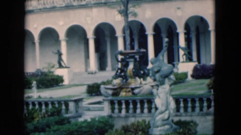 1975 Florida garden view with an ornate fountain, foreground statue, arched colonnade, balustrade, and trimmed plantings.