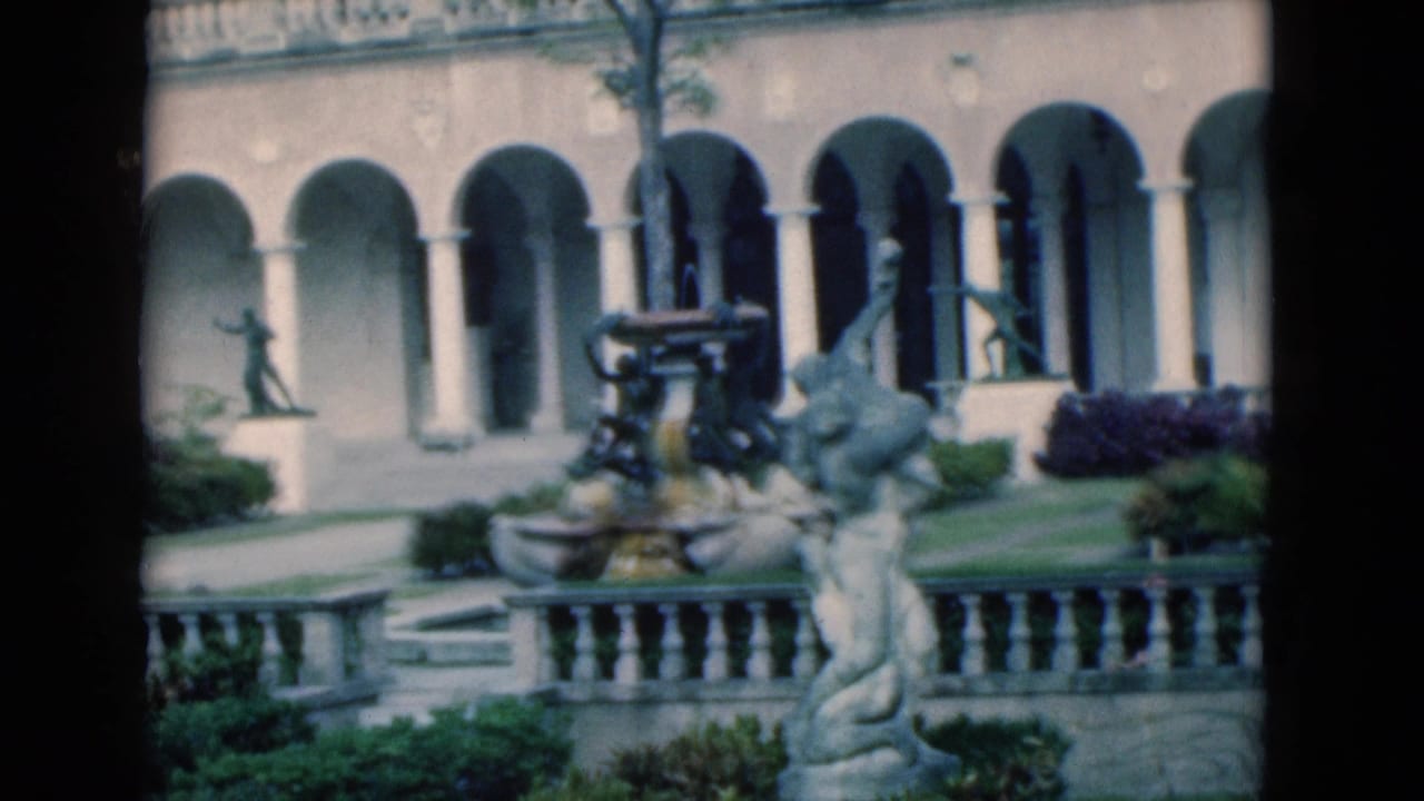 Arched courtyard facade with fountain, sculpture, balustrade, lawn, and shrubs.