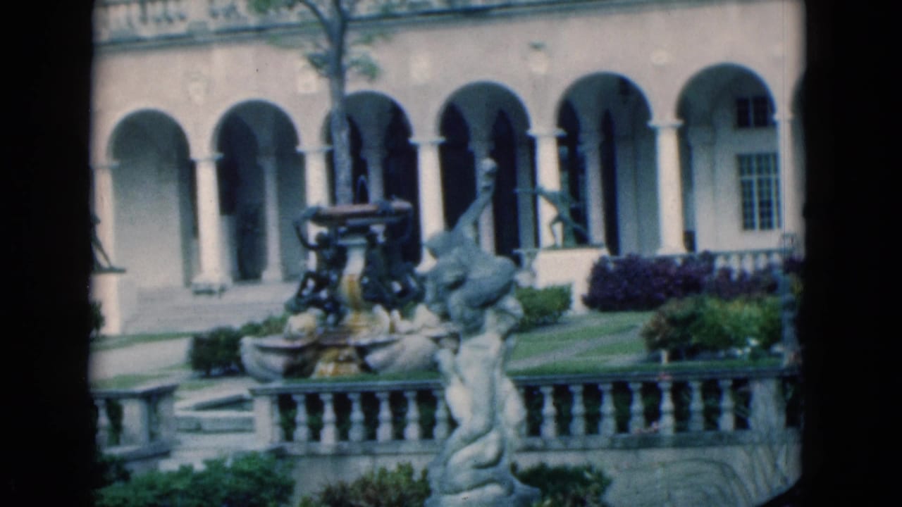 Courtyard fountain and sculpture before a pale arched building and garden beds.