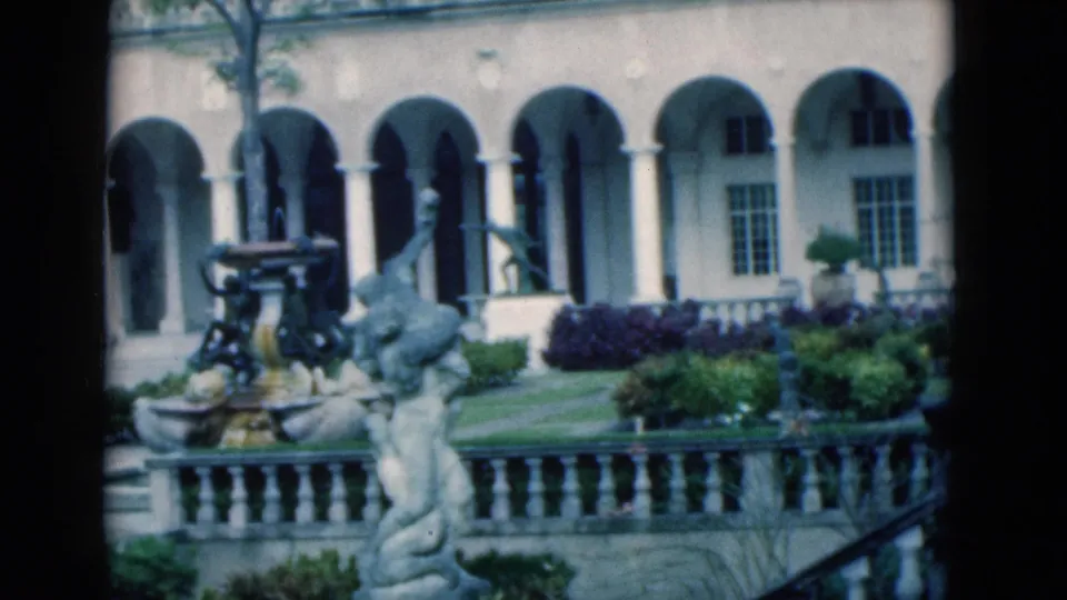 1975 Florida 8mm frame showing a closer view of the courtyard fountain, sculptural figure, arched walkway, windows, and stone balustrade.
