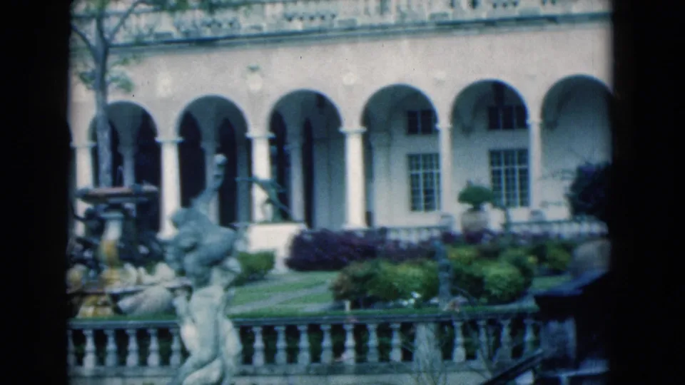 1975 Florida home-movie frame with the arched courtyard facade, partial fountain, sculpture, lawn, shrubs, and stone balustrade in soft focus.