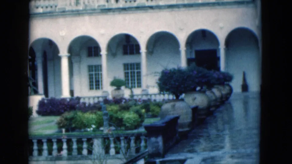 1975 Florida 8mm frame with arched colonnades, windows, stone balustrade, purple shrubs, and formal courtyard plantings.